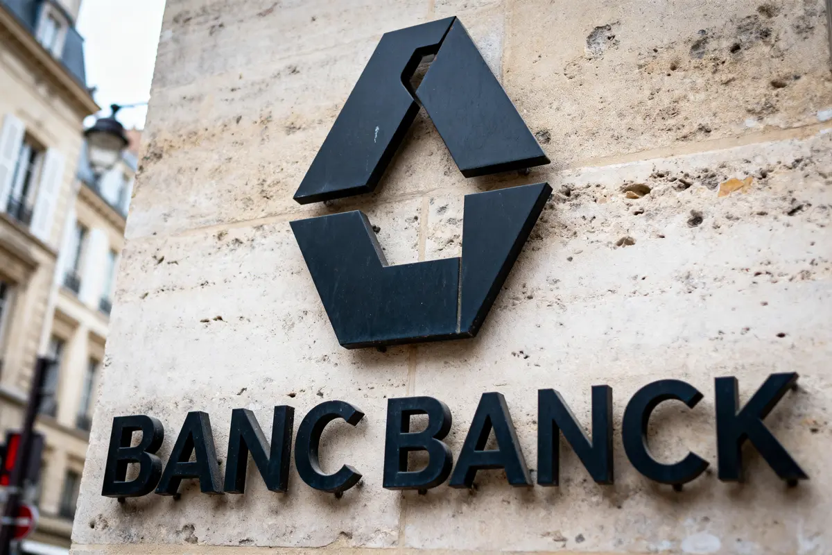 Logo noir géométrique et texte "BANC BANCK" sur mur en pierre. Bâtiments flous en arrière-plan.