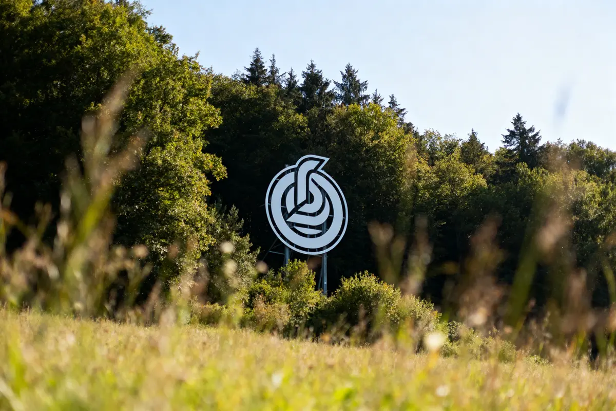 Grand logo blanc circulaire devant forêt verdoyante, ciel bleu, herbe au premier plan floue.