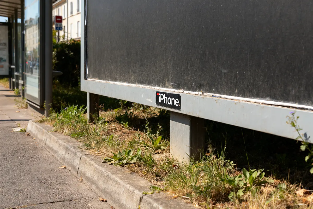 Arrêt de bus avec panneau publicitaire vide, étiquette "iPhone" en bas, herbe autour.