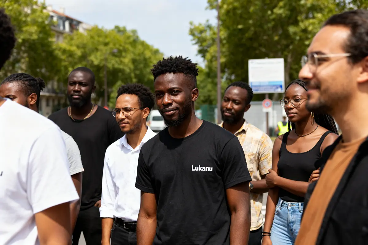 Un groupe de personnes se tenant dehors avec un homme portant un t-shirt Lukanu